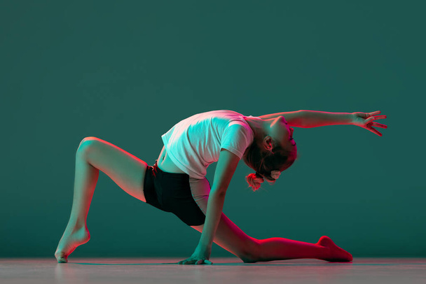 Klein flexibel meisje, ritmische gymnastiek kunstenaar training geïsoleerd op groene studio achtergrond in neon roze licht. Genade in beweging, actie. Oefeningen doen in flexibiliteit. - Foto, afbeelding