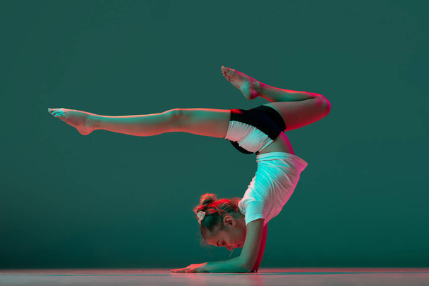 Handstand. Little flexible girl, rhythmic gymnastics artist training isolated on green studio background in neon pink light. Grace in motion, action. Doing exercises in flexibility. - Foto, imagen