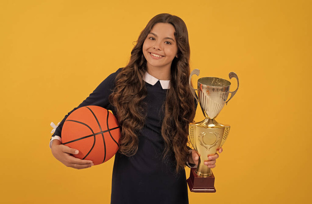 χαρούμενο κορίτσι έφηβος κατέχουν μπάλα μπάσκετ και κύπελλο πρωταθλητής στο κίτρινο φόντο, πρωταθλητής - Φωτογραφία, εικόνα