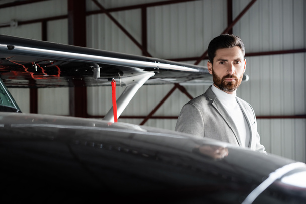 hombre de negocios barbudo de traje mirando la cámara cerca de helicóptero en el estacionamiento  - Foto, Imagen