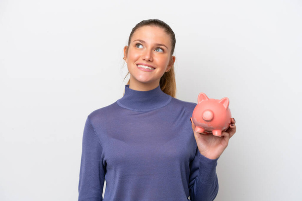 Young caucasian woman holding a piggybank isolated on white background looking up while smiling - Foto, imagen