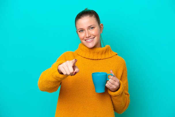 Молода біла жінка тримає чашку ізольовано на синьому фоні пальцем на вас з впевненим виразом
 - Фото, зображення