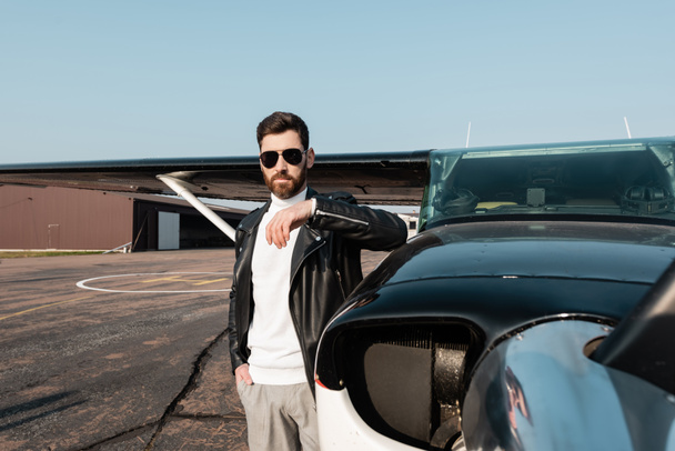 bearded pilot in leather jacket and sunglasses standing with hand in pocket near aircraft  - Foto, imagen
