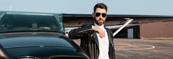 bearded pilot in leather jacket and sunglasses standing near helicopter, banner  - Fotó, kép