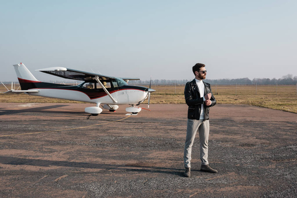 full length of stylish pilot in leather jacket and sunglasses standing near helicopter  - Zdjęcie, obraz