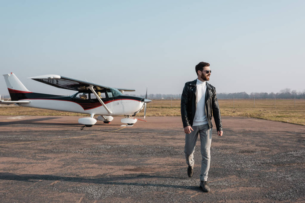 volledige lengte van de stijlvolle piloot in leren jas en zonnebril wandelen in de buurt van helikopter - Foto, afbeelding