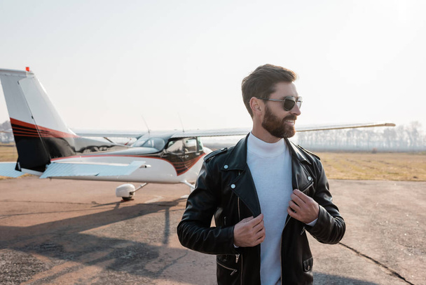 happy pilot in sunglasses adjusting stylish leather jacket  near helicopter - Fotografie, Obrázek