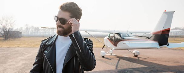 pleased pilot in stylish leather jacket adjusting sunglasses near helicopter, banner - Foto, Bild