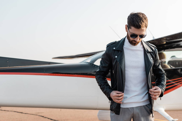 bearded pilot in stylish sunglasses adjusting leather jacket near aircraft  - Фото, зображення