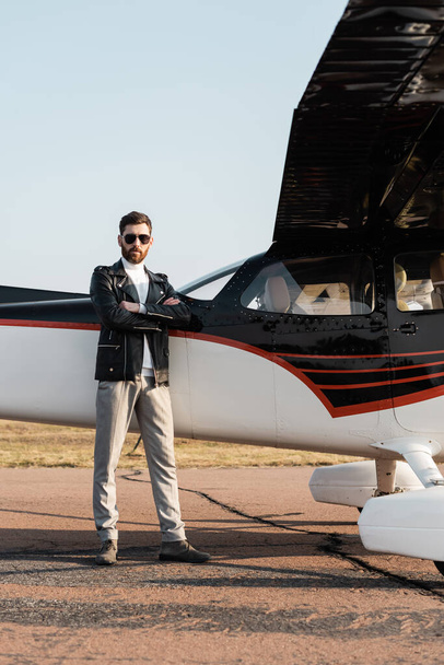 volledige lengte van de bebaarde piloot in stijlvolle zonnebril en leren jas staande met gekruiste armen in de buurt van vliegtuigvleugel  - Foto, afbeelding