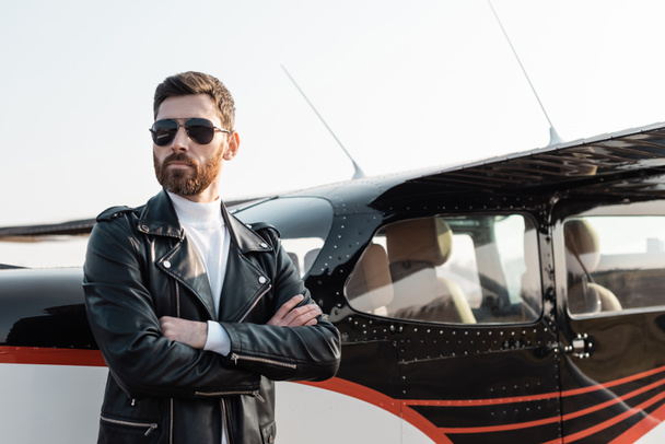 bearded pilot in sunglasses and leather jacket standing with crossed arms near helicopter  - Fotografie, Obrázek
