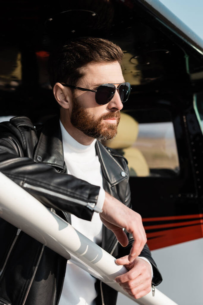 confident bearded man in sunglasses and black leather jacket near helicopter - Foto, Imagem