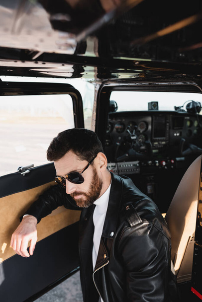 bearded pilot in stylish sunglasses and black leather jacket near opened door of helicopter - 写真・画像