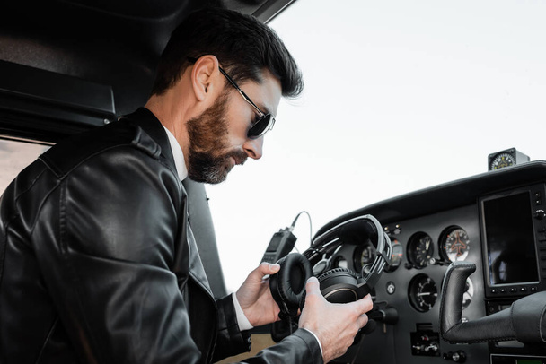 piloto barbudo em óculos de sol e jaqueta de couro preto segurando fones de ouvido no cockpit de helicóptero  - Foto, Imagem
