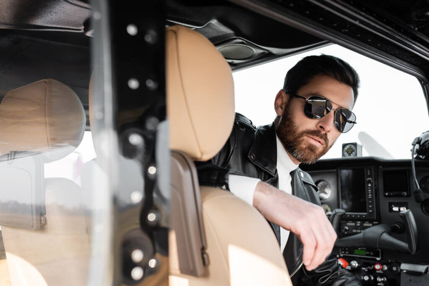 bearded pilot in stylish sunglasses and leather jacket sitting in cockpit of helicopter  - Foto, Imagem