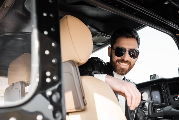 piloto positivo en gafas de sol con estilo y chaqueta de cuero sentado en la cabina de helicóptero  - Foto, imagen