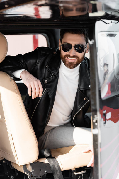 cheerful pilot in stylish sunglasses and leather jacket smiling while sitting in cockpit of helicopter  - Photo, image