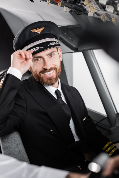 piloto barbudo na tampa de ajuste uniforme e sorrindo perto de co-piloto borrado  - Foto, Imagem