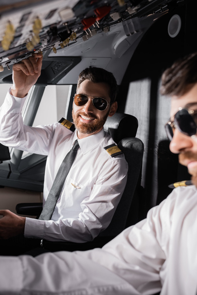 piloto alegre em óculos de sol atingindo painel aéreo perto co-piloto no simulador de avião - Foto, Imagem