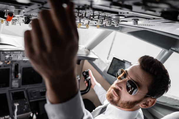 high angle view of bearded pilot in sunglasses reaching overhead panel in plane simulator - Photo, Image