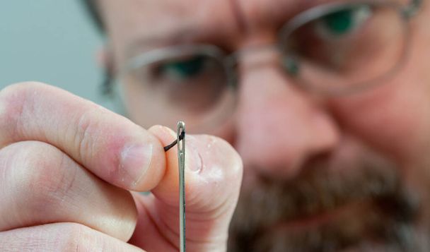 man's face wearing glasses (blurred in background) sticks the thread into the needle (foreground) - Foto, Imagem