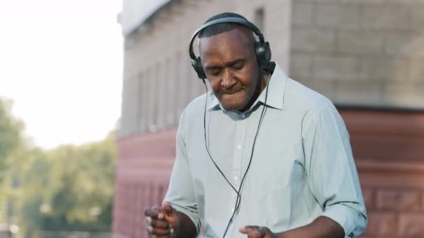 Opgewonden gelukkige oudere man met een draadloze hoofdtelefoon die plezier heeft muziek luisteren in smartphone, dans, vrolijke persoon met pensioen leeftijd in moderne oortelefoons doen alsof ze buiten drummen - Video