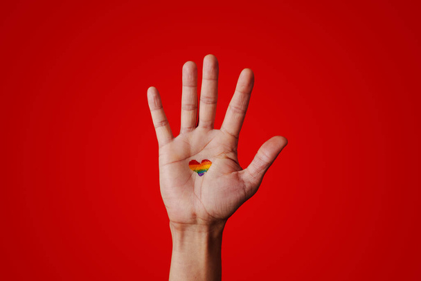 la mano levantada de un hombre con una bandera de arco iris en forma de corazón pintada en su palma sobre un fondo rojo - Foto, imagen