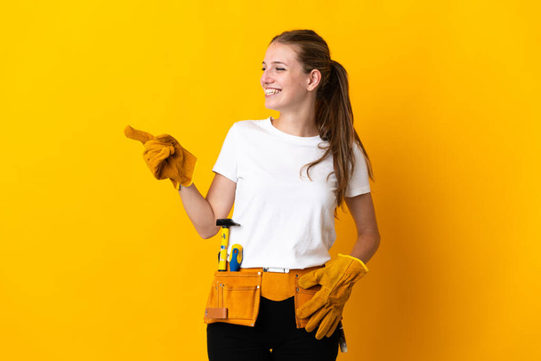 Young electrician woman isolated on yellow background pointing finger to the side and presenting a product - 写真・画像