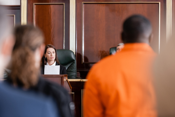 aanklager leest rechtszaak in de buurt van rechter en beschuldigde Afro-Amerikaanse man op wazig voorgrond - Foto, afbeelding