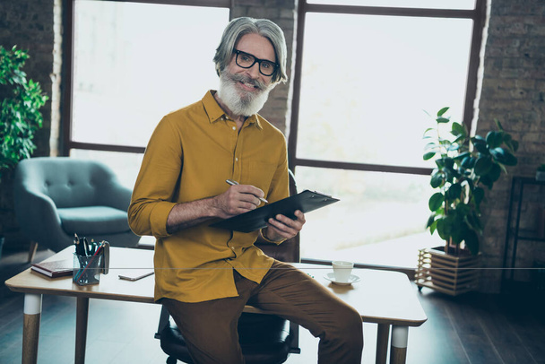 Photo of cheerful mature guy dressed yellow shirt spectacles writing clip board indoors workplace workstation - Foto, Imagen