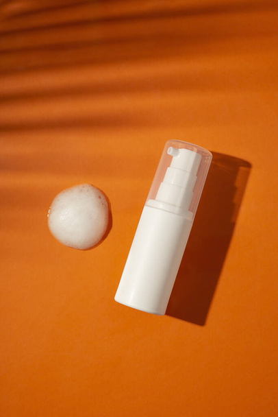 Plastic white bottle with pump and the appearance of the texture of the foam on an orange background. Skincare cleansing cosmetic in top view with beautiful palm shadows. Beauty concept for face body care - Photo, Image