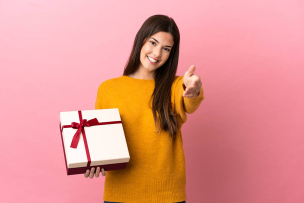Teenager Brazilian girl holding a gift over isolated pink background shaking hands for closing a good deal - Photo, Image