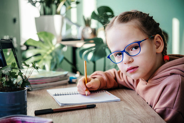 Primary school girl in eyeglasses writing in her notebook while having online lesson during covid lockdown, distance education for children - Фото, изображение