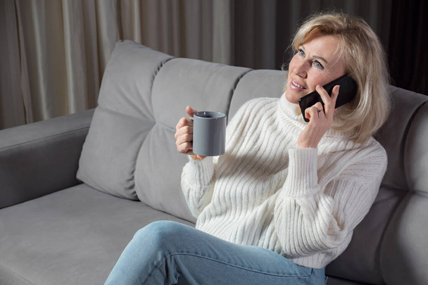 Blonde-haired woman talks via smartphone and smiles on sofa - Photo, Image