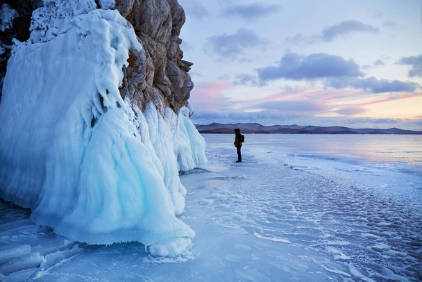 Озеро Байкал на заході сонця. Людина подорожує по кризі озера. Турист дивиться на бурульки на скелях..  - Фото, зображення