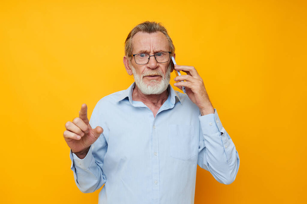 old man in a blue shirt and glasses talking on the phone yellow background - Φωτογραφία, εικόνα