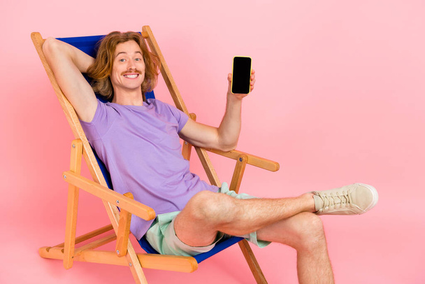 Portrait of attractive cheerful guy sitting in chair holding in hand device display touchscreen isolated over pink pastel color background. - 写真・画像
