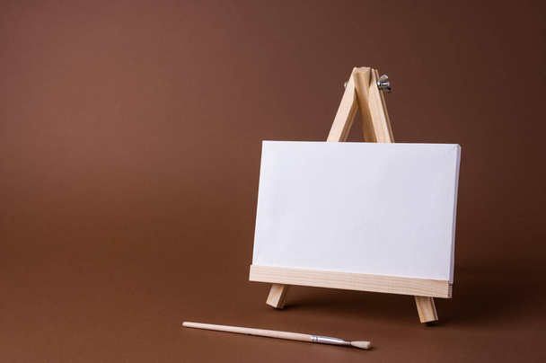 White canvas on a small wooden easel stand minimal concept on the brown backdrop. Empty blank mock up frame for design with copy space. - Zdjęcie, obraz