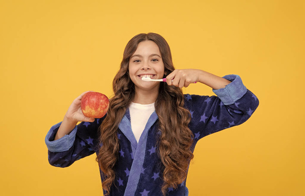 joyeuse adolescente avec les cheveux bouclés en pyjama confortable brossant les dents et tenir pomme, dentisterie - Photo, image