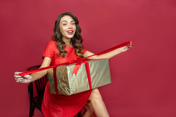 Joyful brunette woman with a gift in red dress on a red background - Photo, Image