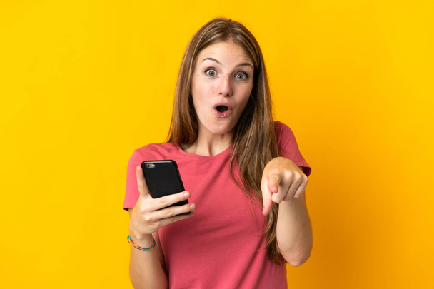 Молода жінка за допомогою мобільного телефону ізольована на жовтому тлі здивована і спрямована спереду
 - Фото, зображення