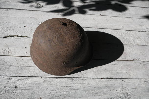 Erinnert euch an den Krieg! Ein rostiger deutscher Helm aus dem Zweiten Weltkrieg, durchbohrt von einer Kugel, auf einem von der Sonne erleuchteten weißen Tisch - Foto, Bild