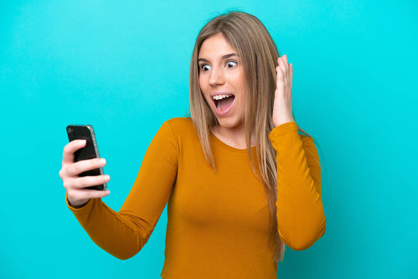 Young caucasian woman isolated on blue background looking at the camera while using the mobile with surprised expression - Foto, afbeelding