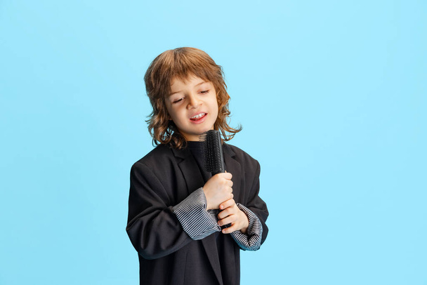 Studio shot of liitle cute boy, kid in image of stylish hairdresser, barber isolated on blue background. Concept of occupation, emotions, childhood, studying - Zdjęcie, obraz