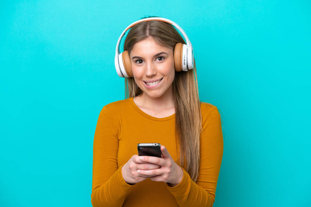 Jeune femme caucasienne isolée sur fond bleu écoutant de la musique avec un mobile et regardant devant - Photo, image