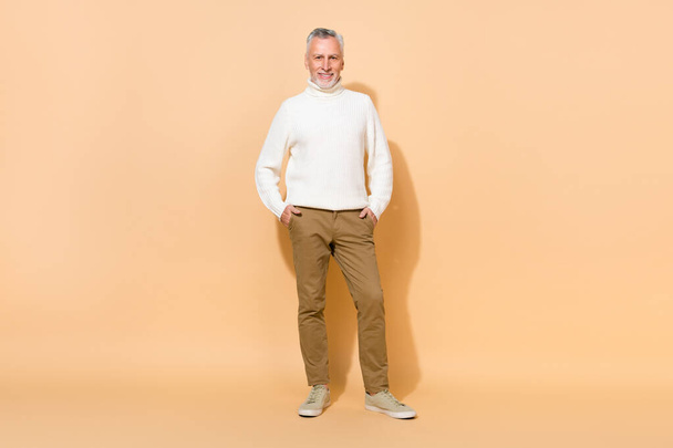 Full length body size view of attractive cheerful grey-haired man holding hands in pockets isolated over beige pastel color background - Valokuva, kuva