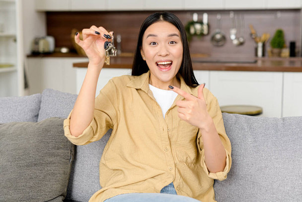 Relocation concept. Headshot of excited female moving in new home or apartment. Cheerful asian woman homeowner looking at camera and points finger at the keys - Fotó, kép