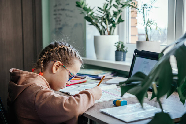 Menina da escola primária em óculos escrevendo em seu caderno enquanto tem aula on-line durante o confinamento vívido, educação à distância para crianças - Foto, Imagem