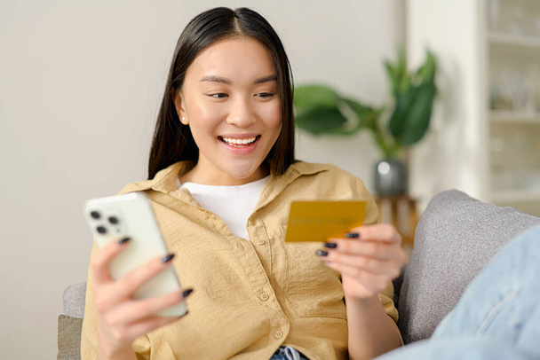 Vrolijke jonge Aziatische vrouw met behulp van smartphone winkelen app voor het kopen van online, met een creditcard en input bankgegevens, zit op de bank - Foto, afbeelding