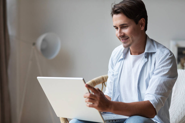 Portrait of a young smiling man with a laptop indoors. Chat with friends online. - 写真・画像